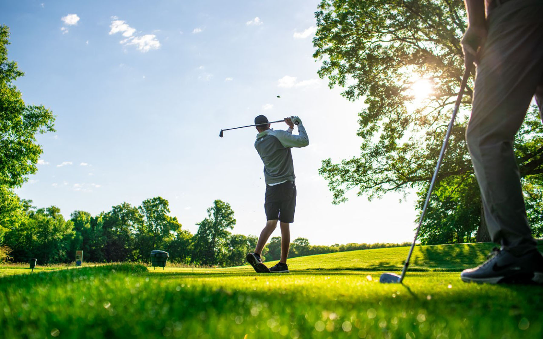 Two people enjoy a lush, green golf course in the Spring Guide. One swings their club under the sunlit trees, while the other stands in the foreground, ready for their turn on this perfect spring day.