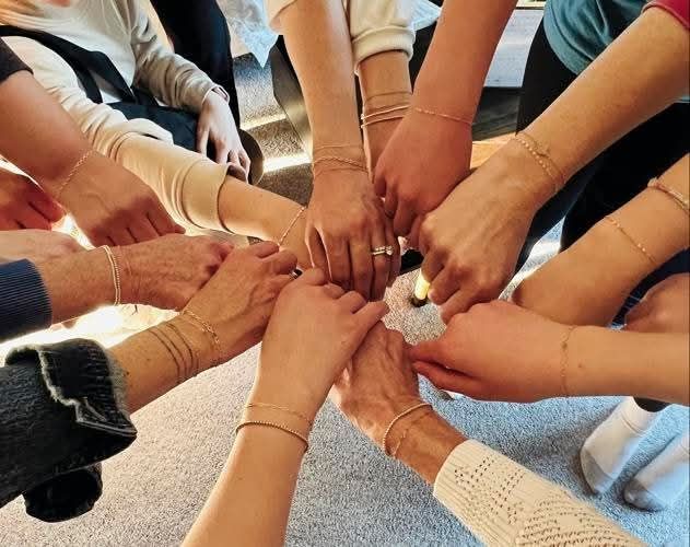 A group of people stand in a circle, each extending one hand to the center, showing matching gold chain bracelets from a Jewelry Pop Up on their wrists, symbolizing unity and togetherness.