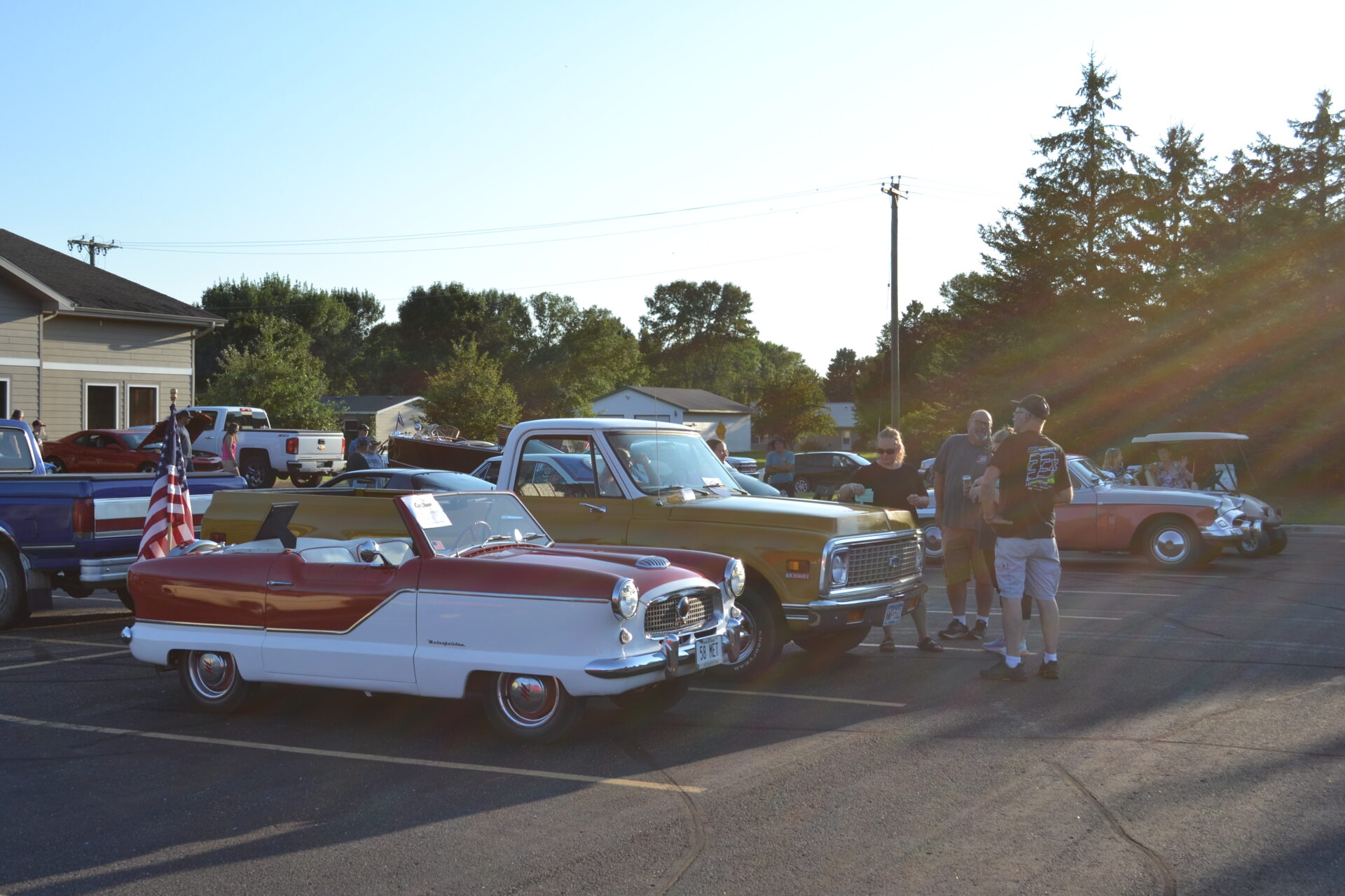 A group of people stands near vintage cars, including a red and white convertible with an American flag, during the sunny Brandon Car Show, with more classic vehicles and trees in the background.