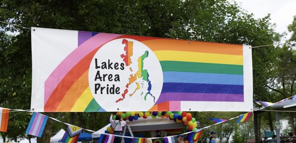 A banner reading "Lakes Area Pride" with a rainbow and map graphic hangs outdoors at this lively Pride in the Park. Rainbow flags and colorful balloons decorate the area, with trees and people attending the LGBTQ event visible in the background.