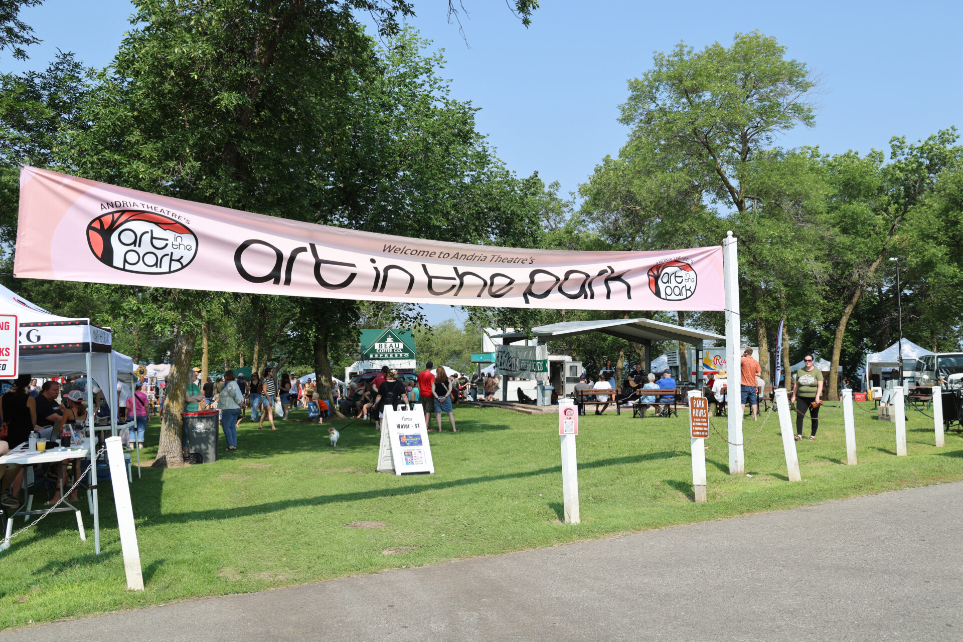 A large “Art in the Park” banner hangs over a grassy area as people stroll among colorful art booths and tents at an outdoor festival surrounded by trees on a sunny day.