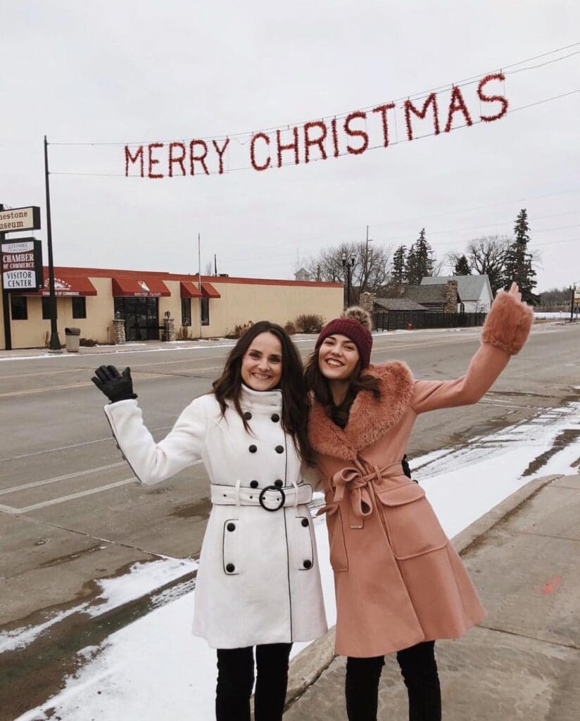 Two women in winter coats smile and wave on a snowy street under a festive "Merry Christmas" banner, capturing the joyful holiday spirit. The overcast sky, buildings, and trees set a cozy winter scene in the background.