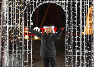A young child in a dark coat stands under an archway decorated with white holiday lights at night, smiling and reaching out both arms joyfully—ready to explore Alexandria, MN. Colorful lights twinkle warmly in the blurred background.