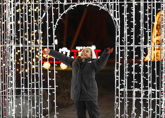 A young child in a dark coat stands under an archway decorated with white holiday lights at night, smiling and reaching out both arms joyfully—ready to explore Alexandria, MN. Colorful lights twinkle warmly in the blurred background.