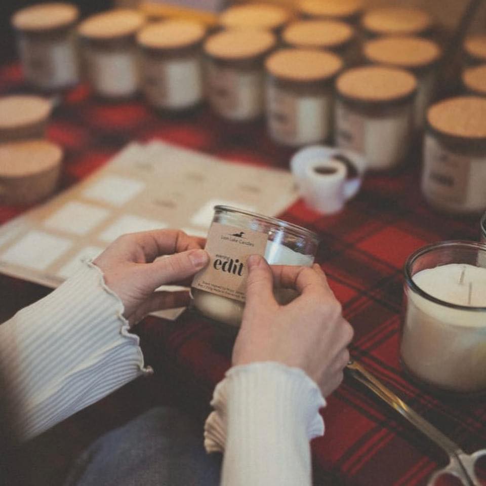 A person holds a glass jar candle labeled "edit" over a red plaid table covered with other candles and Candle Making Workshop supplies.
