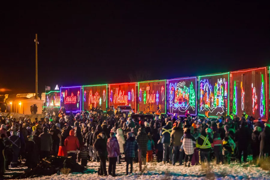 A large crowd gathers at night in front of the brightly lit Holiday Train 2025, decorated with colorful Christmas lights and festive designs, with snow covering the ground.