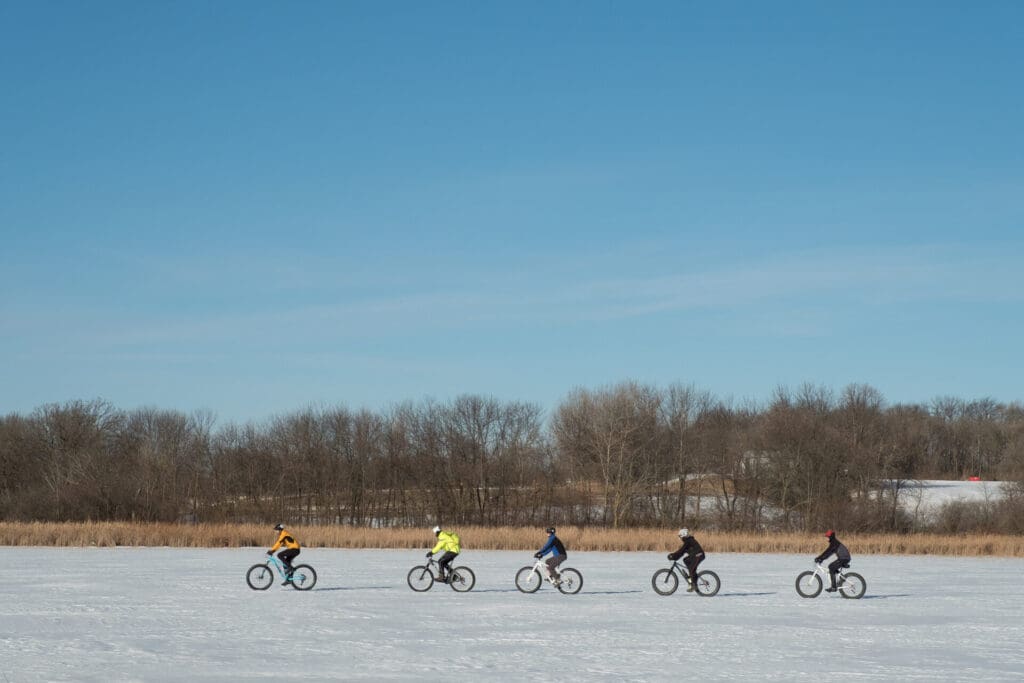 Five people riding bicycles in a single line across a snowy, open field near Alexandria MN, with bare trees and blue sky in the background—perfect for those who rent outdoor equipment for winter adventures.