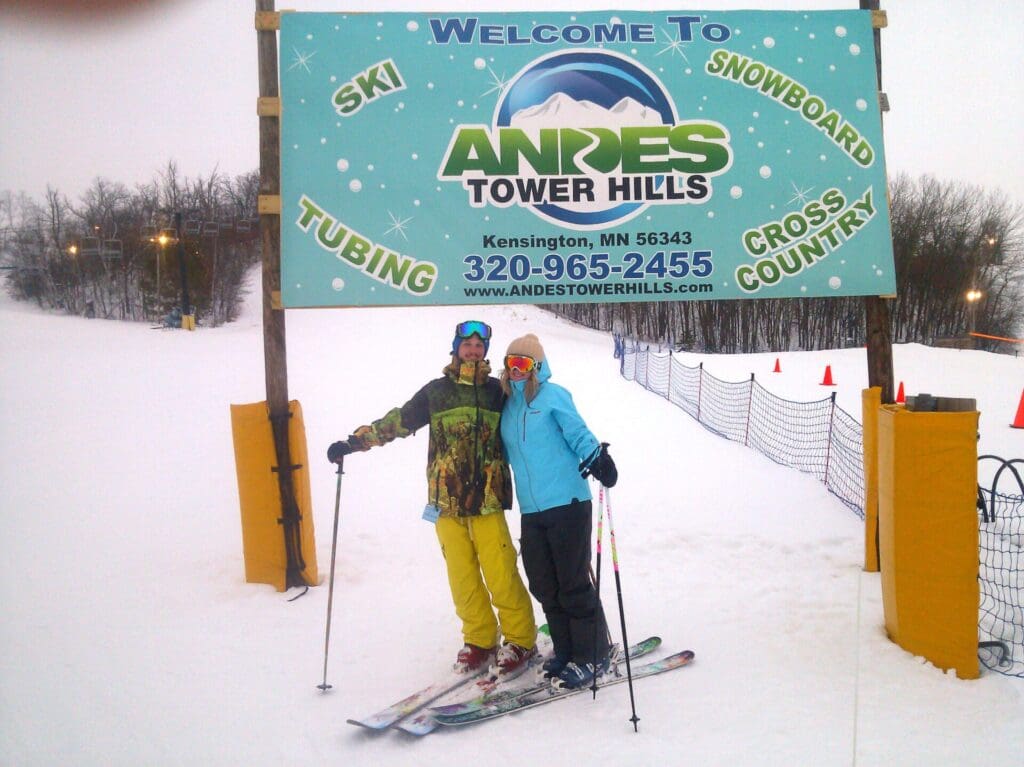 Two people in ski gear stand on snow under a large Andes Tower Hills sign near Alexandria MN, advertising skiing, snowboarding, tubing, and cross-country fun. Trees and snowy landscape are visible in the background. Outdoor gear rental available.