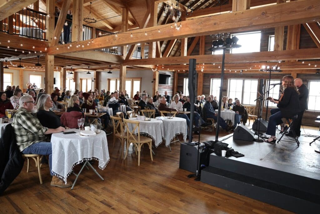 A large crowd sits at round tables with white tablecloths, watching two musicians perform on a small stage in a rustic wooden hall with exposed beams and string lights during one of the lively Alexandria winter festivals.