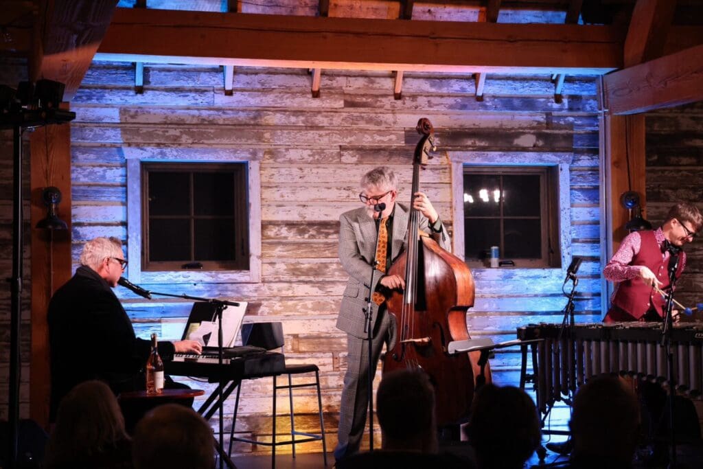 Three musicians perform on stage in a rustic, wood-paneled room during the Alexandria Winter Festivals; one plays keyboard and sings, one plays upright bass, and one plays vibraphone. Warm lighting highlights the scene as an audience looks on.