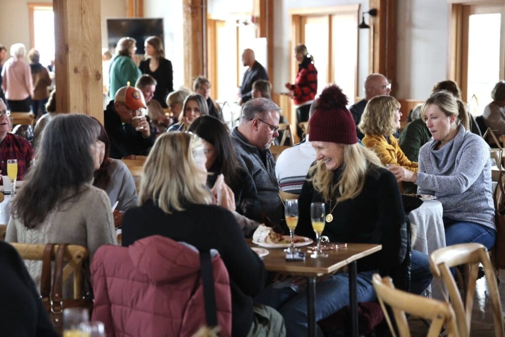 A lively restaurant scene during Alexandria Winter Festivals, with people of various ages sitting at wooden tables, eating, drinking, and chatting. Warm sunlight filters in through large windows, creating a cozy, inviting atmosphere.
