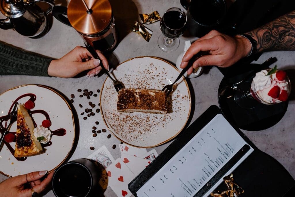 Two people share a dessert at a table in Alexandria, set with coffee, wine, cheesecake with berries, scattered coffee beans, and playing cards; one arm bears a tattoo—perfect for a cozy Valentine's Day.