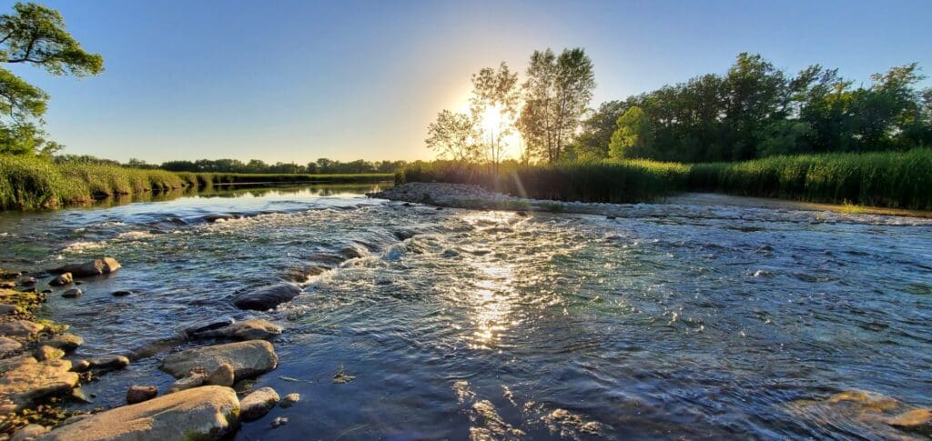 A shallow river flows over rocks under a clear blue sky in Alexandria MN, with the sun shining brightly behind tall trees and lush green vegetation along the banks—a perfect Spring Break escape.