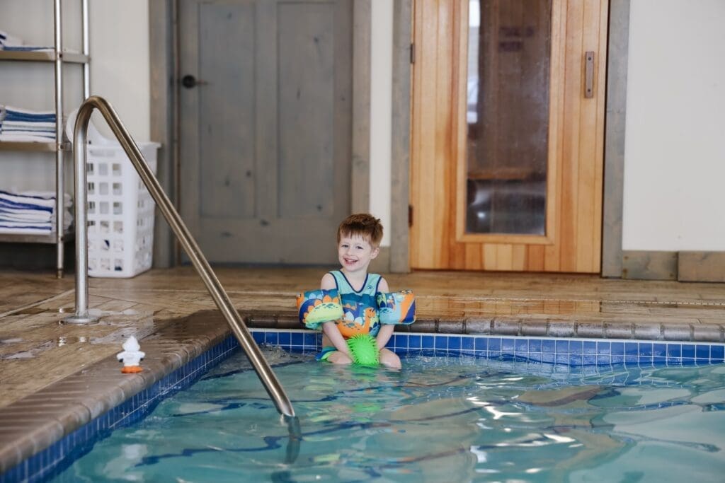 A young boy wearing colorful floaties sits on the edge of an indoor pool, smiling. Behind him are a wooden door, a sauna, tiled floor, and a stack of towels—perfect for Spring Break fun in Alexandria MN.