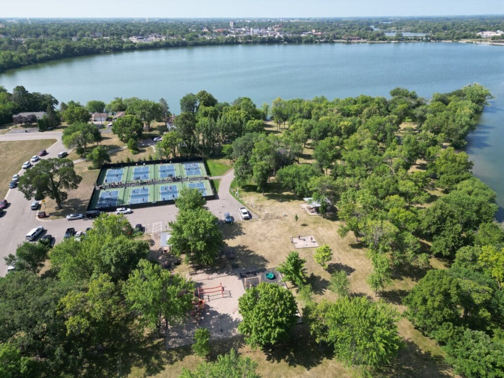 Aerial view of a lakeside park in Alexandria MN with tennis courts, a playground, and trees. Cars are parked along the road, and the lake is visible in the background under a clear sky—perfect for your Spring Break travel guide.