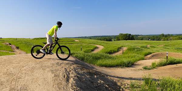 A person wearing a helmet and neon shirt rides a bike on a dirt trail surrounded by green grass and rolling hills under a clear blue sky, enjoying the fresh air of Spring Break near Alexandria MN.