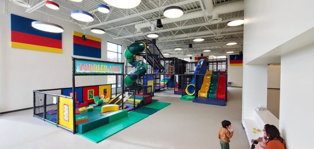 Indoor playground in Alexandria MN with colorful climbing structures, slides, and a fenced toddler area. A child stands near an adult at the counter; the bright space features large windows and modern decor—perfect for your Spring Guide adventure.
