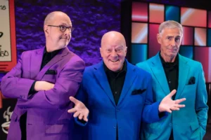 Three men stand together smiling in brightly colored suits—purple, blue, and turquoise—against a vibrant, illuminated backdrop. The cheerful trio looks ready to perform Triple Espresso, bringing their live show energy to Theatre L'Homme Dieu.