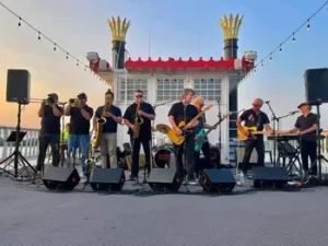 The Belfast Cowboys, a band of eight musicians, performs outdoors on a stage with brass, guitars, and keyboards. Decorative smokestacks and string lights glow behind them against the backdrop of a stunning sunset sky.