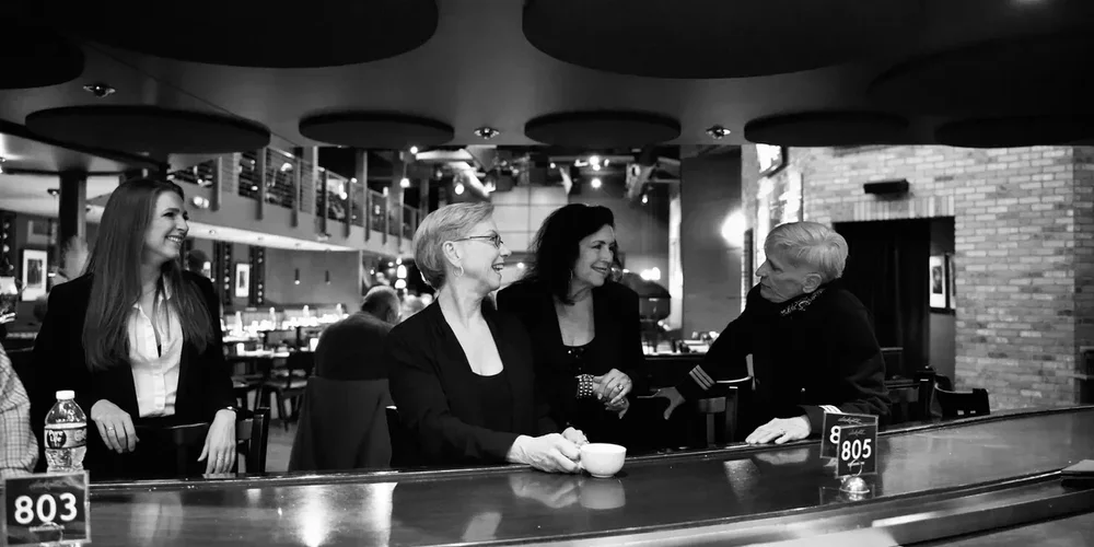Four women sit and smile together at a curved bar counter in a modern restaurant or lounge, enjoying pleasant conversation about Songs by Boys by Girls. The scene is captured in timeless black and white.