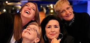 Four women are close together, smiling and laughing, in a warmly lit indoor setting. The joyful mood feels like the soundtrack to favorite songs—just like "Boys by Girls," it's all about friendship and happiness.