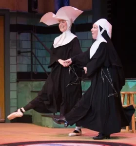 Two women dressed as nuns, one in a large winged white headdress, joyfully dance together on stage at Theatre L'Homme Dieu, holding hands and lifting a leg in the air—a lively moment from the musical comedy Nunsense.