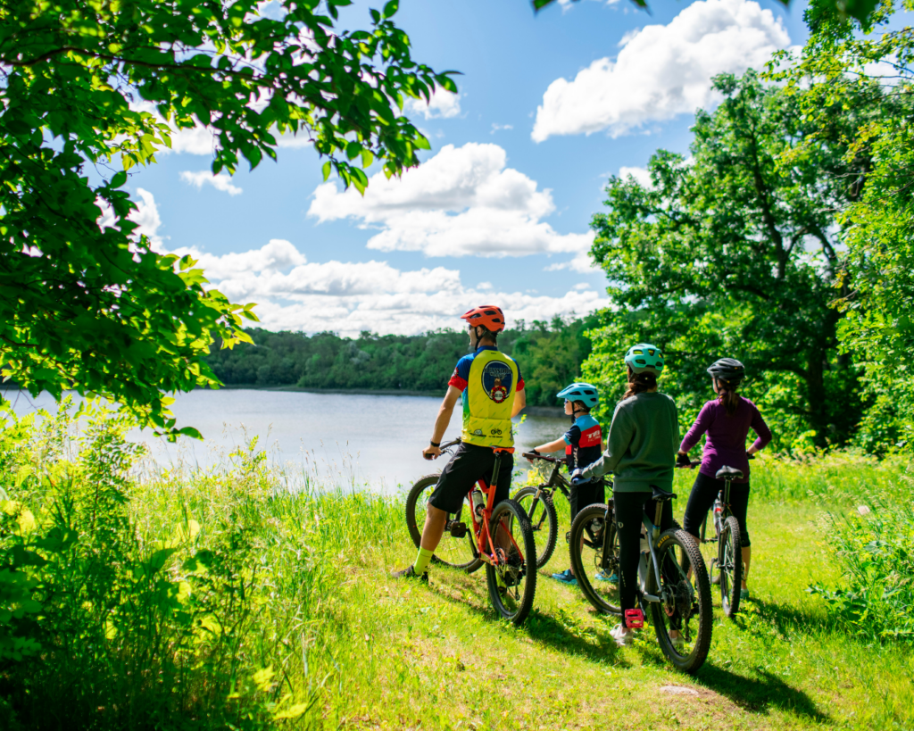 Four people wearing helmets and colorful clothes pause on their bikes near a lake in Alexandria, surrounded by lush green trees and grass under a sunny, blue sky with scattered clouds on Memorial Day Weekend.