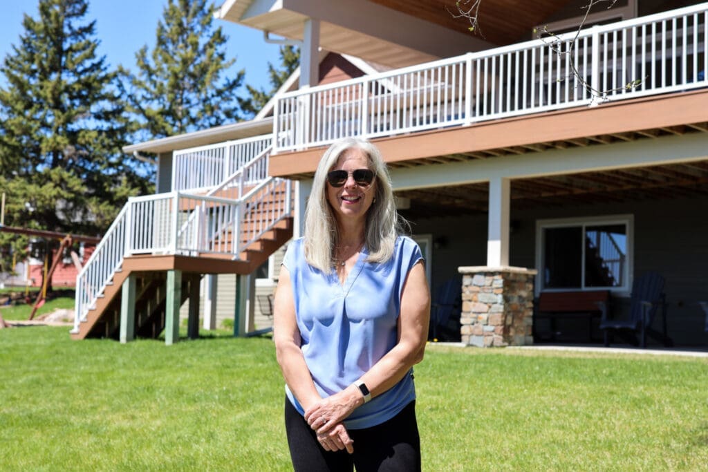 A woman with long gray hair, wearing sunglasses and a light blue sleeveless top, stands smiling on vibrant green grass in front of a house with a large porch and white railings on a sunny day.