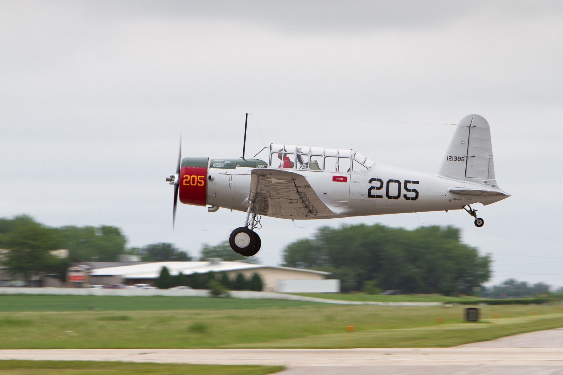 A vintage single-engine military aircraft marked "205" takes off from the runway at the Wings Over Alexandria Air Show 2026, with green grass, buildings, and trees visible in the background under a cloudy sky.
