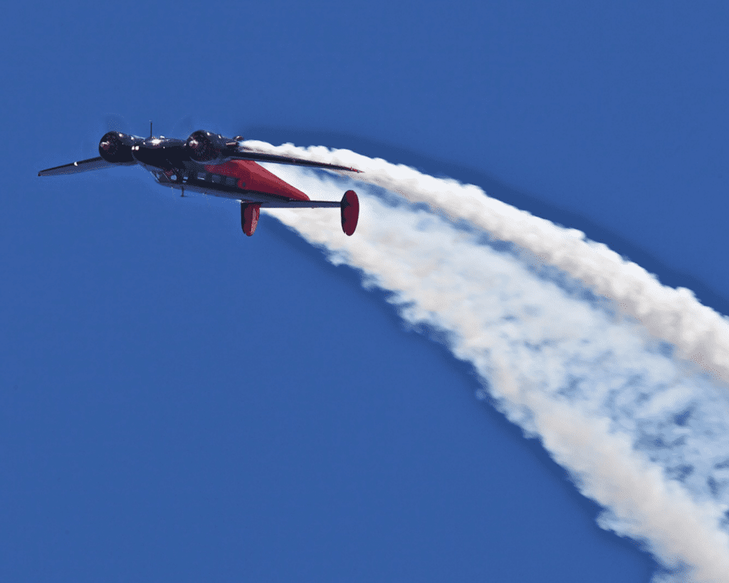 An airplane with a red underside flies through a clear blue sky, leaving a thick white smoke trail behind it.