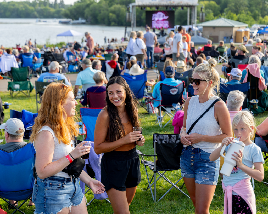 Three women and a young girl stand and chat, smiling, at an outdoor event with a crowd seated in lawn chairs near a lake. A stage and screen are visible in the background. The weather is sunny and warm.