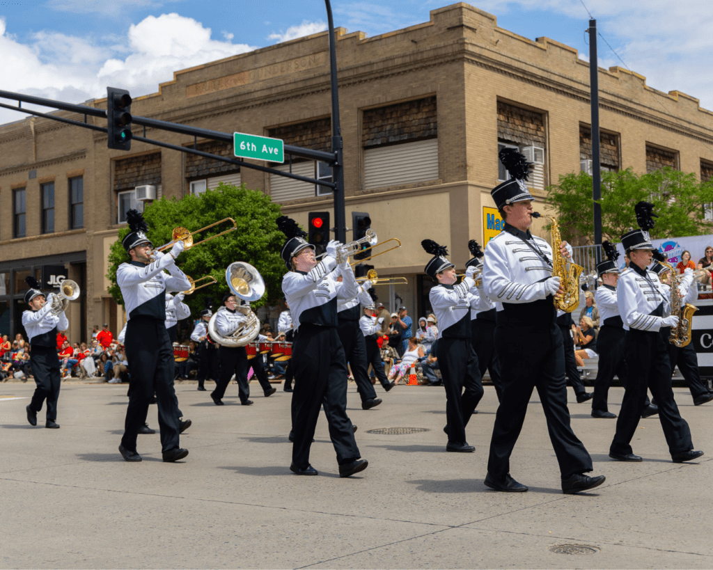 A marching band in black and white uniforms plays brass instruments while walking in a parade on a city street, with spectators sitting along the sidewalk and a large tan brick building in the background.