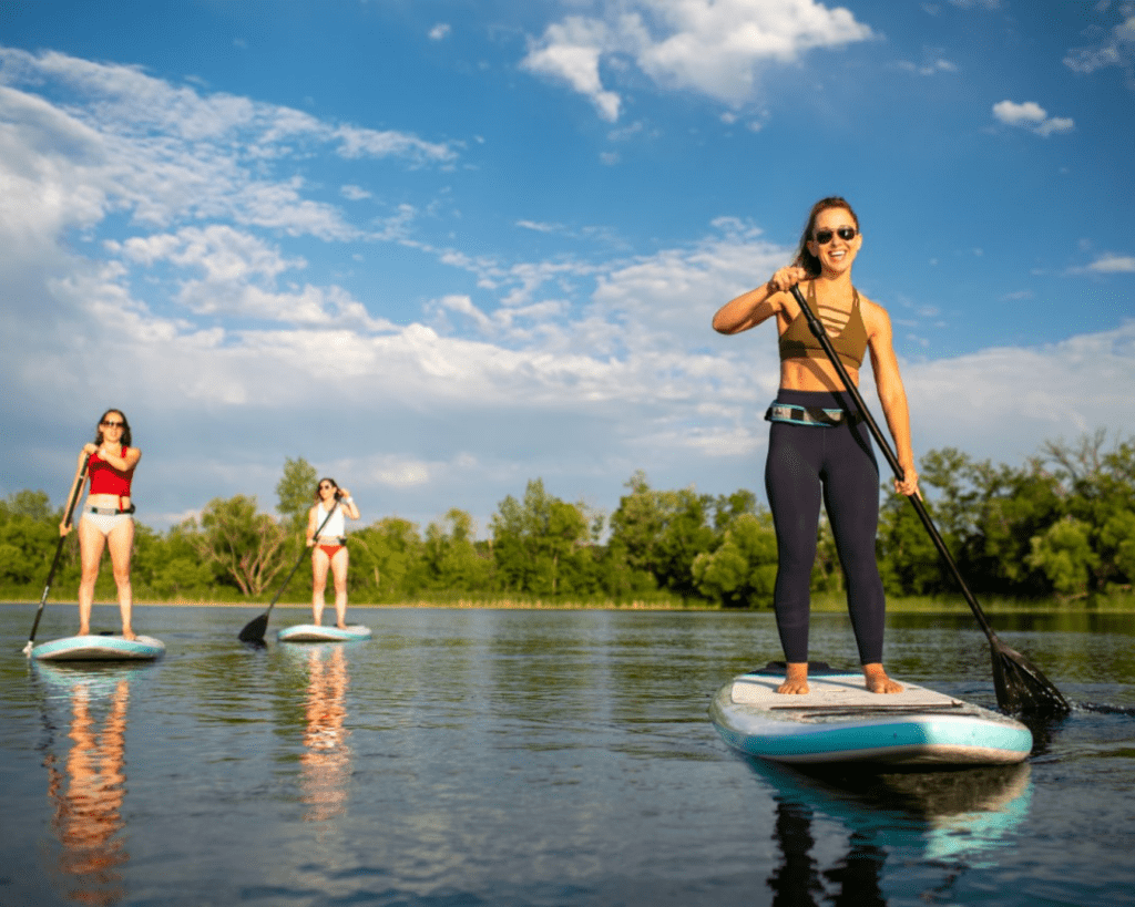 Three women are paddleboarding on a calm lake under a blue sky with scattered clouds, surrounded by green trees. One woman in front is smiling and wearing sunglasses.