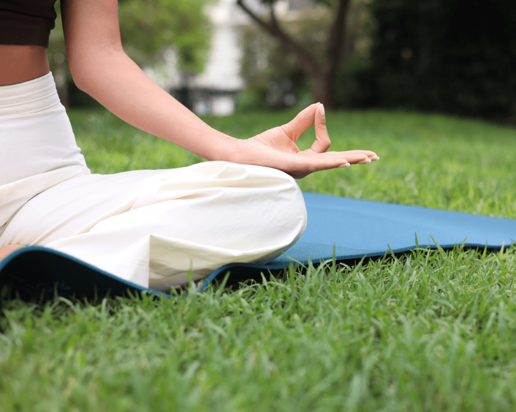 A person sitting cross-legged on a blue yoga mat outdoors on green grass, with their hand resting on their knee in a meditation pose. The background is blurred with trees and foliage.
