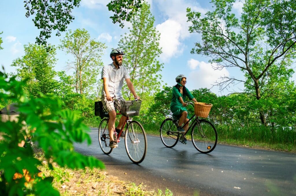 Two people wearing helmets ride bicycles with baskets along a paved road, surrounded by green trees and foliage in Lake Country, savoring the season on a sunny day—a perfect entry for any Summer Bucket List.