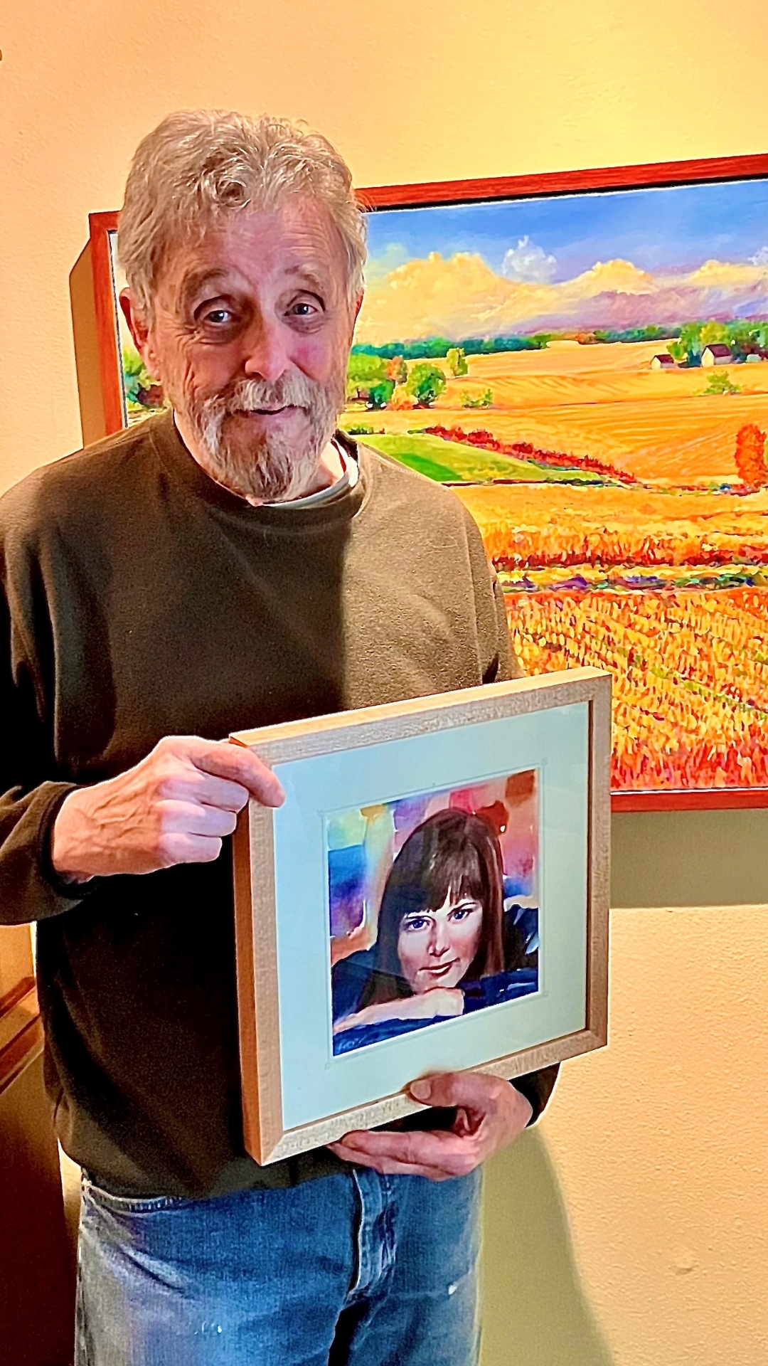 An older man with a gray beard and mustache stands indoors, smiling and holding a framed portrait of a woman. Behind him is a colorful painting of a bright, rural landscape—an inspiring setting for our Artist of the Month.