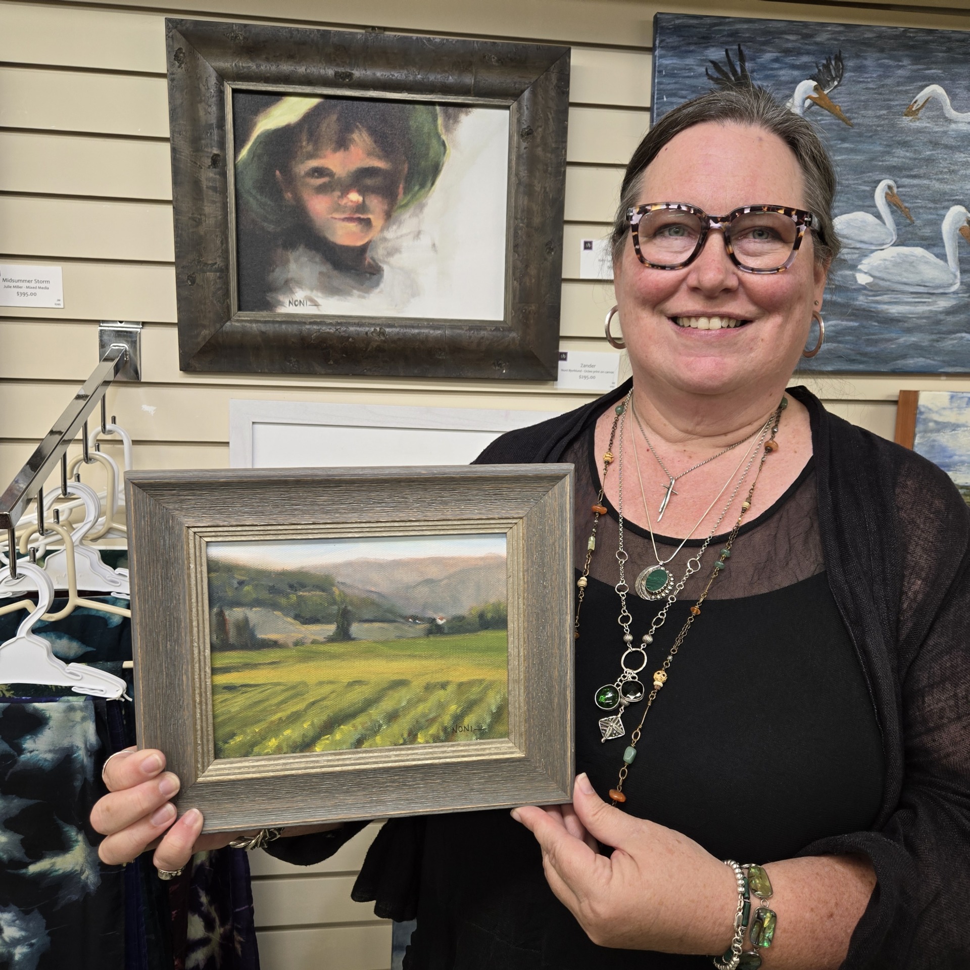 A smiling woman, Naomi Bjorklund—Artist of the Month at Alexandria Art Guild—holds a framed landscape painting in an art gallery. Behind her are other artworks, including a portrait of a child and a painting of swans.
