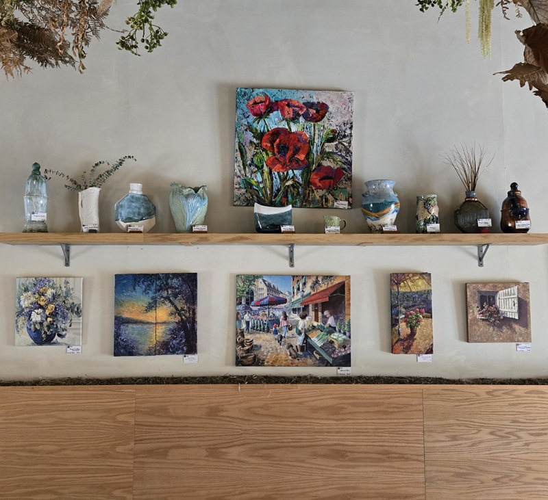 A wall display features colorful paintings, including landscapes inspired by the Lakes of Minnesota, with pottery and vases arranged on a wooden shelf above the artwork. The light, minimalistic background enhances the vibrant scenes.