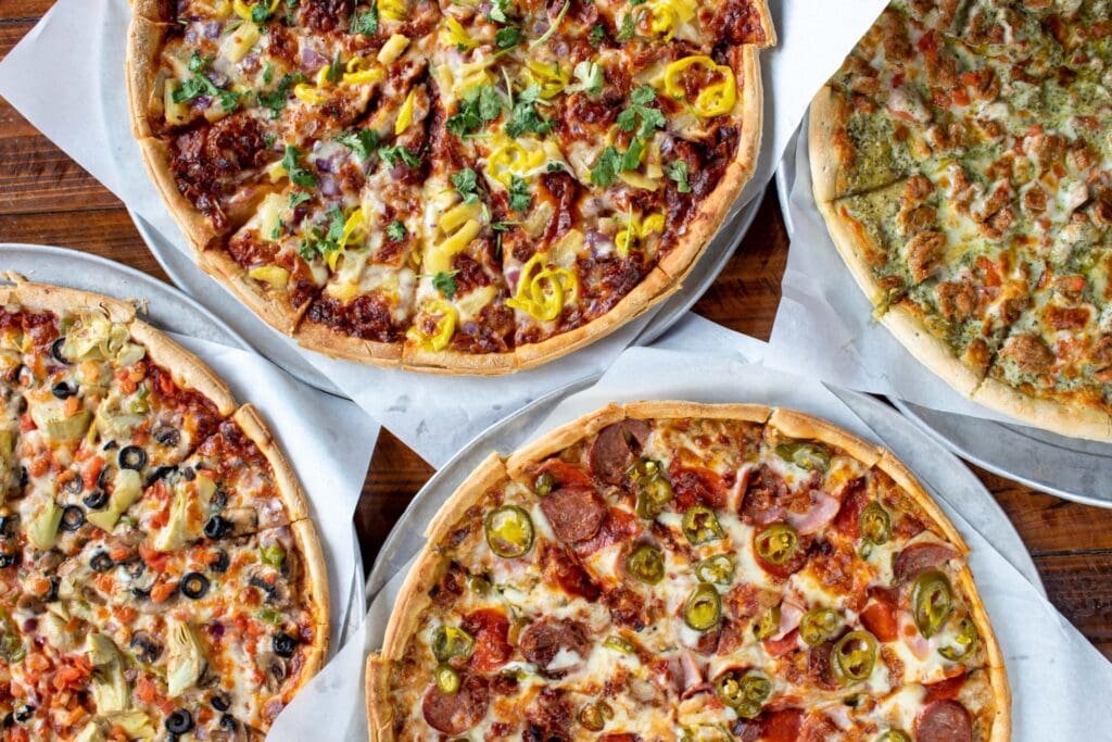 Four pizzas with different toppings, including pepperoni and jalapeños, artichoke and olives, banana peppers and cilantro, and a green herb pizza, arranged on parchment paper on a wooden table.