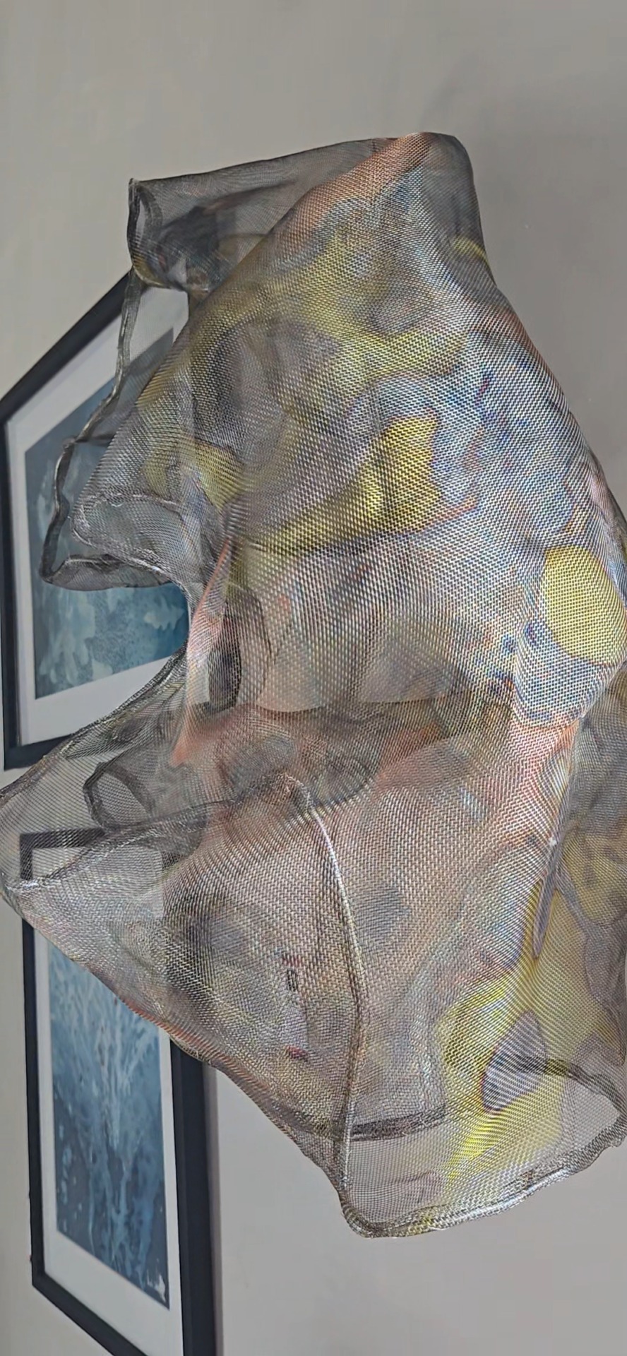 A semi-transparent, mesh-like fabric with yellow and gray hues is draped in front of two framed blue art prints on a wall, creating an evocative display reminiscent of installations discussed in recent Artist Talk sessions hosted by the Alexandria Art Guild.