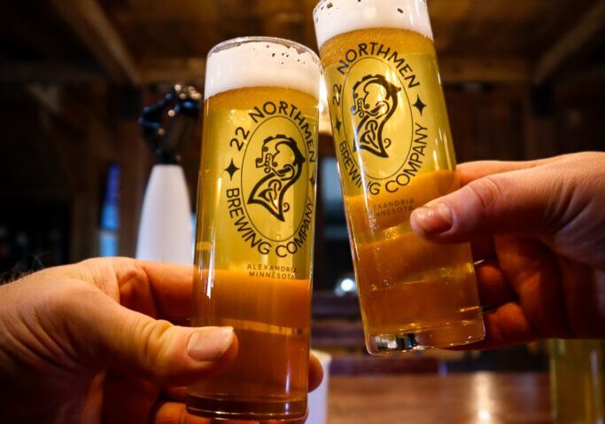 Two hands clink tall beer glasses with "22 Northmen Brewing Company" logos, stoking the spirit of the brewery against a rustic wooden backdrop. Both glasses are filled with golden beer and crowned with a frothy head.