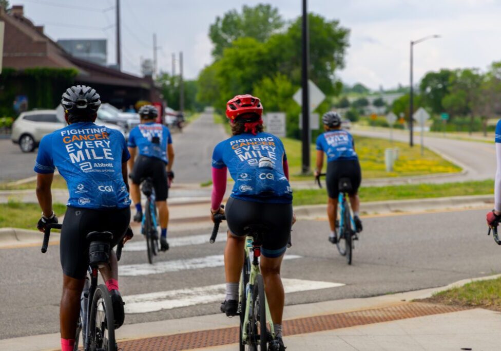 Five cyclists in matching blue jerseys reading "FIGHTING CANCER EVERY MILE" ride together on a cloudy day, heading toward a crosswalk. They're part of the Alexandria to Texas to Alaska bike ride, raising awareness as they journey onward.