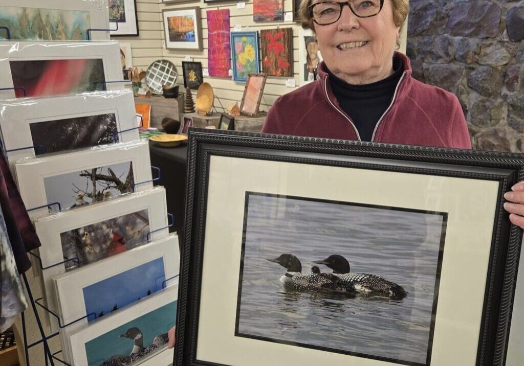 A smiling woman with glasses holds a framed nature photography print of two loons on the water, standing in an art shop surrounded by various framed artworks and photo cards, celebrating Photography Day.