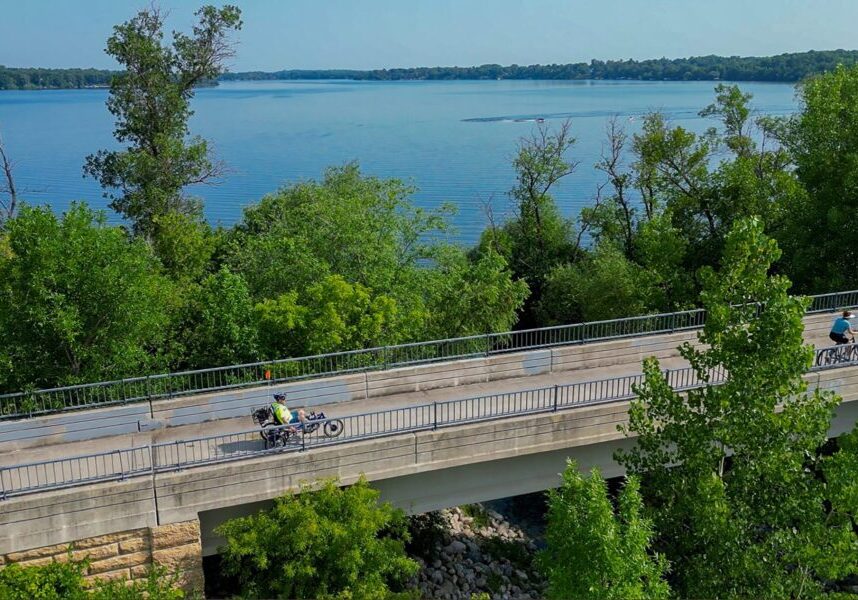 Two people ride bicycles on the Central Lakes Trail, crossing a bridge surrounded by lush green trees, with a large blue lake and forested shoreline in the background under a clear sky.