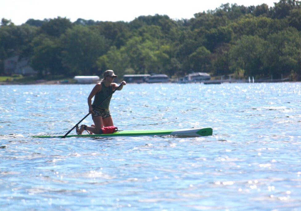 A person wearing a cap and sunglasses kneels on a paddleboard, paddling across sunlit Lake Ida with trees and boats visible in the background.