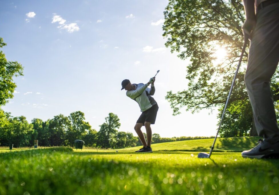 A golfer is mid-swing on the lush, green course at Geneva Golf Club under a clear sky, with sunlight streaming through the trees. Another person stands nearby, watching.