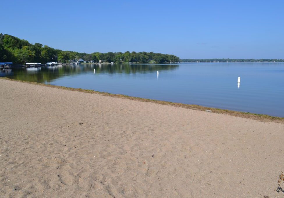 A sandy beach with calm water, a tree-lined shoreline, and a clear blue sky awaits at Lake Le Homme Dieu Beach in Alexandria, MN. White buoys float in the water and a few docks with boats are visible in the distance.