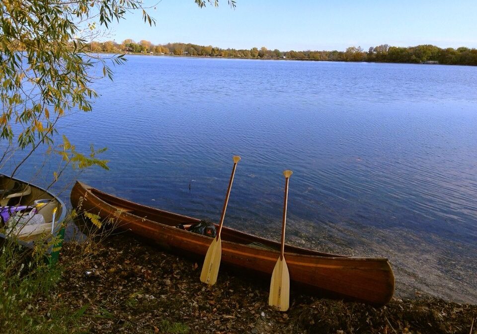 A wooden canoe rests on the shore of Lake Brophy, with two paddles propped against it. Trees frame the scene, and the water stretches toward a distant tree-lined horizon under a clear blue sky.