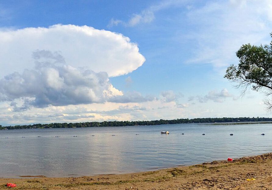 A sandy beach leads to the calm waters of Lake Darling, with a distant treeline on the horizon. The sky is partly cloudy, featuring a large billowy cloud. A few buoys and a floating object rest on the lake’s surface.