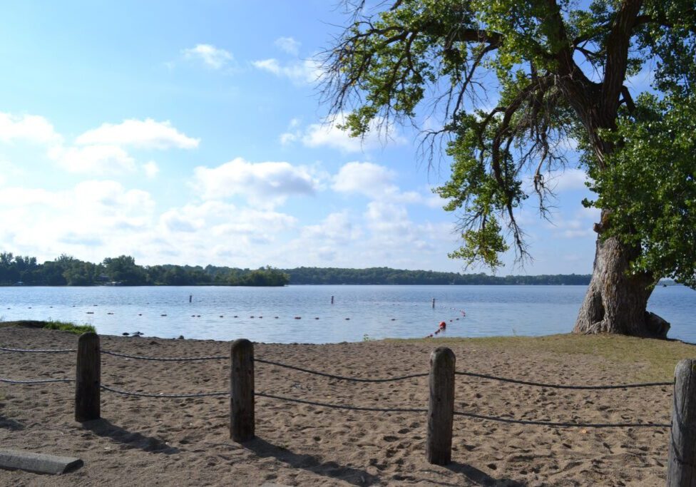 A sandy beach with wooden posts and rope fencing overlooks calm Lake Latoka in Alexandria, Minnesota. A large leafy tree stands on the right, and the sky is partly cloudy with sunlight shining through.